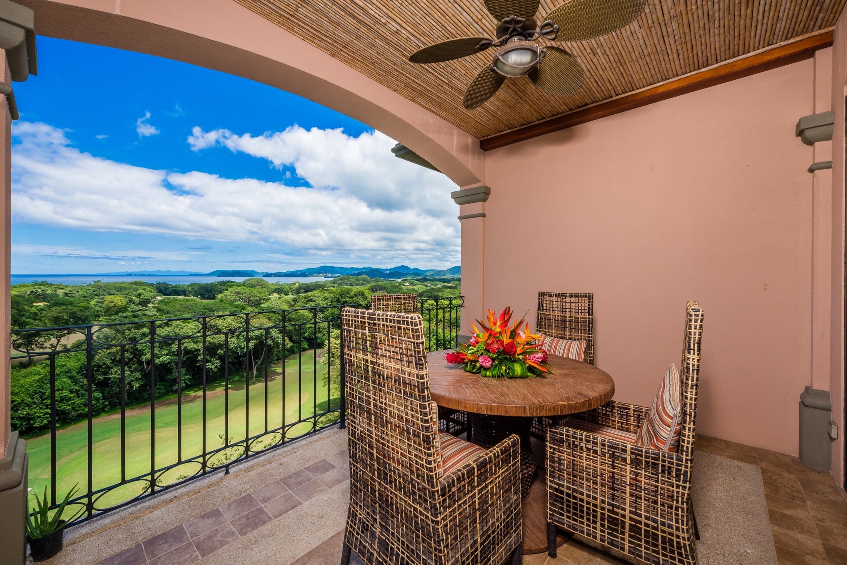 Un balcón con mobiliario de mimbre, una mesa redonda con un centro de mesa floral y vistas a un paisaje verde.