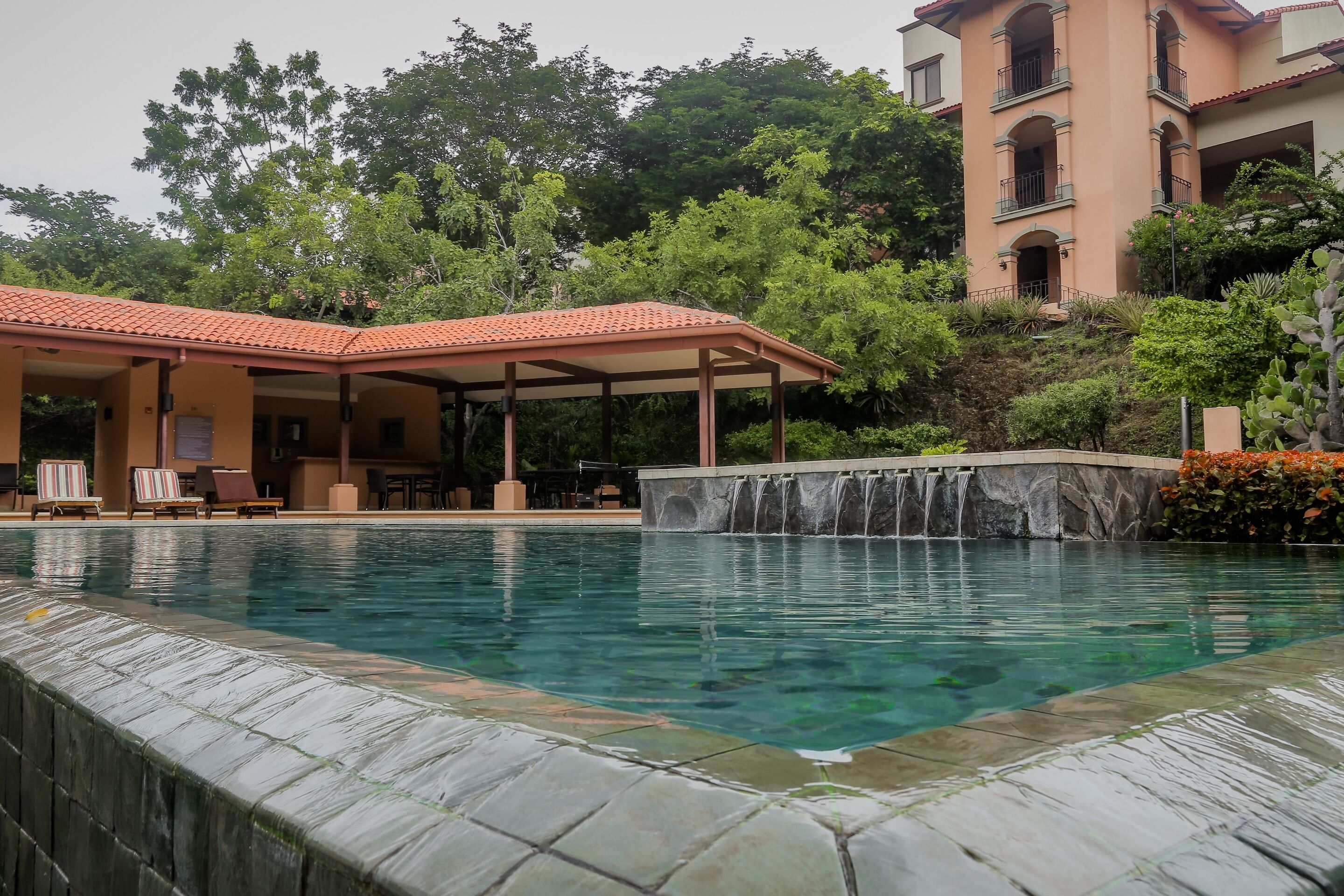A swimming pool with a waterfall feature, surrounded by lounge chairs and a covered seating area.