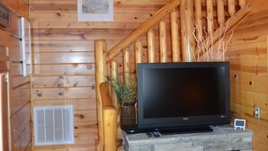 Ein Holzhütten-Interieur mit Kamin aus Stein, einer Holztreppe und einem Flachbildfernseher auf einem Steinkaminbrett.