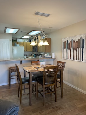 Un comedor con mesa y sillas de madera, una cocina al fondo y una pared con un cuadro de un bote de remos.