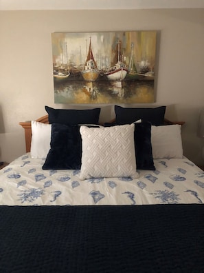 Una cama con una colcha estampada, varios almohadones y un cuadro de barcos en la pared de arriba.