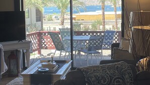 A living room with a TV, a coffee table, and a patterned throw. There is a sliding glass door leading to an outdoor patio with chairs and a table.