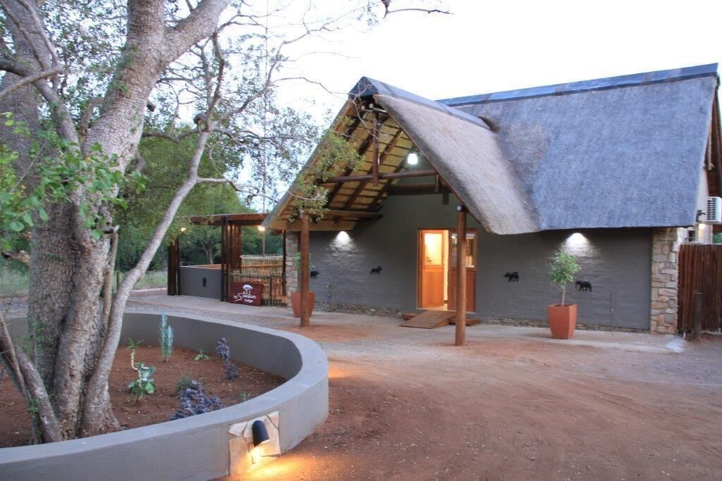 A rustic building with a sloped roof, a concrete planter with plants, and a dirt driveway.