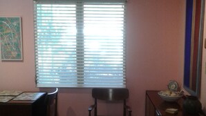 Ein Zimmer mit Esstisch, zwei Stühlen, einem Fenster mit Jalousien, einer Wand mit einem Gemälde und einer kleinen Kommode mit einer Vase.
