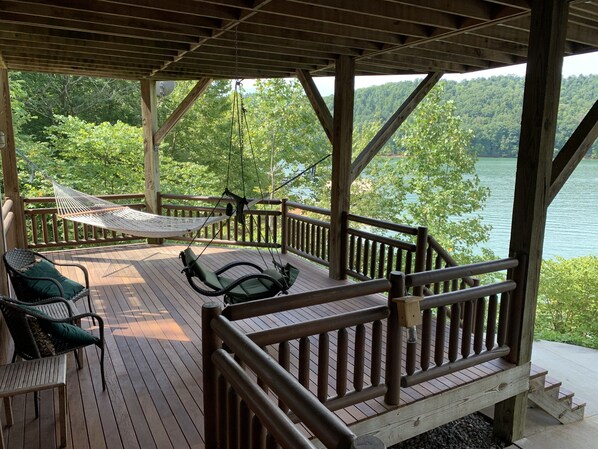 Una terraza de madera con una hamaca, sillas y vistas a un lago rodeado de árboles.