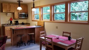 Une cuisine avec des meubles en bois, un four à micro-ondes, une machine à café et une fenêtre donnant sur les arbres. On y trouve une table à manger avec des chaises et un tabouret de bar.