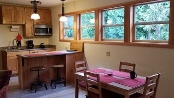 A kitchen with wooden cabinets, a microwave, a coffee maker, and a window with a view of trees. There is a dining table with chairs and a bar stool.