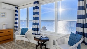 Una habitación con dos ventanas grandes, una silla de mimbre con cojín azul, una mesita redonda con lámpara y un aparador de madera.