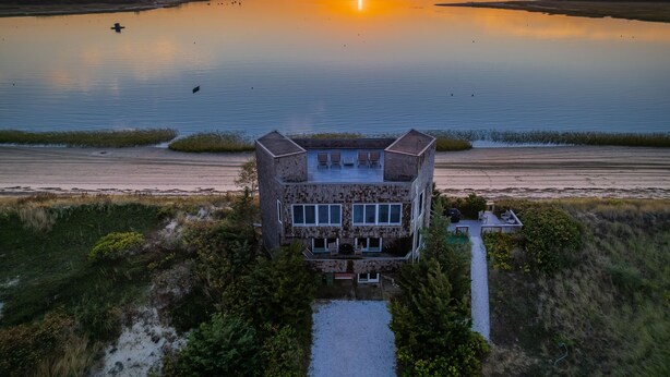 Edificio de dos pisos con techo plano y ventanas amplias, ubicado en una zona verde con vista a una masa de agua tranquila y un atardecer al fondo.