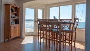 Un comedor con una mesa y sillas de madera, un aparador de madera con cristalería, y ventanas grandes que ofrecen vista al mar.