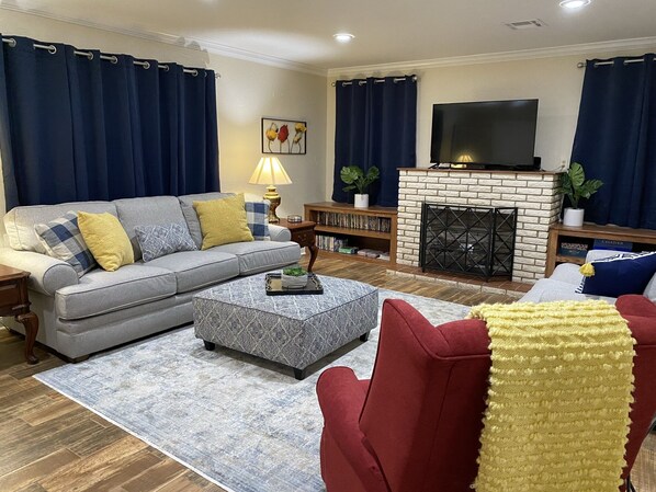 A living room with a grey sofa, red armchair, and patterned ottoman. A flat-screen TV is mounted above a brick fireplace.