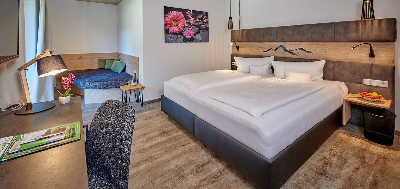 Una habitación de hotel moderna con una cama grande, un escritorio con una lámpara, una mesita con una planta y un cuadro en la pared.