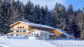 A large wooden chalet with multiple balconies and a snow-covered roof, surrounded by dense forest.