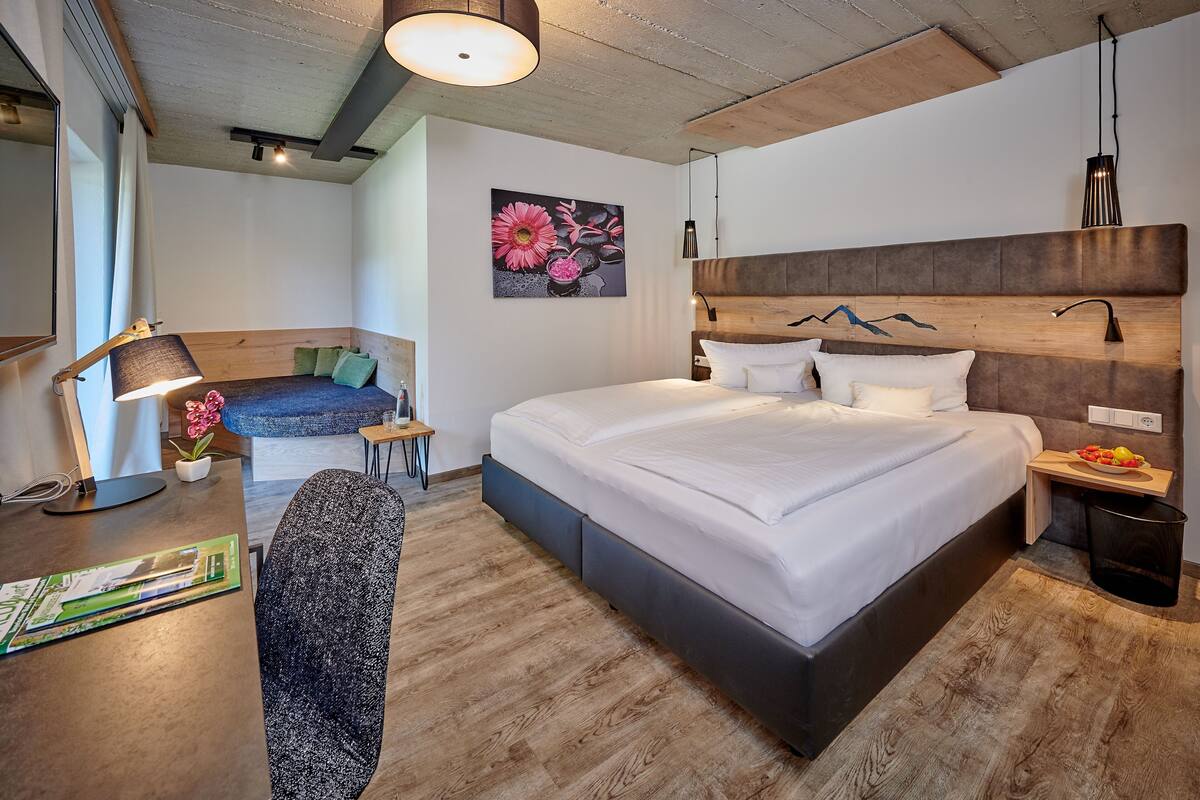 A modern hotel room with a large bed, a desk with a lamp, a small table with a plant, and a painting on the wall.