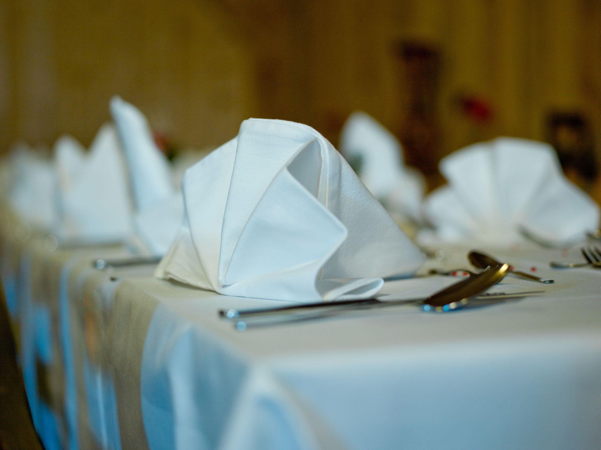 A table set with white napkins folded into a triangular shape, a fork, and a knife on a table covered with a white tablecloth.