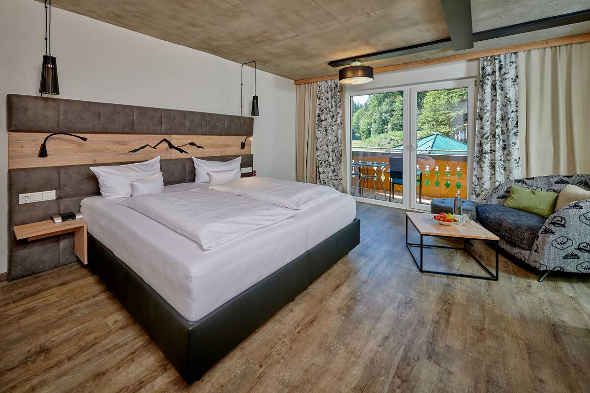 A modern hotel room with a large bed, a sofa, a small table, and a balcony with outdoor seating.