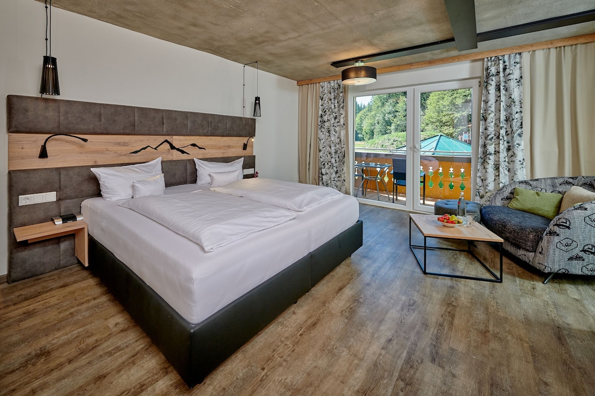 A modern hotel room with a large bed, a sofa, a small table, and a balcony with outdoor seating.