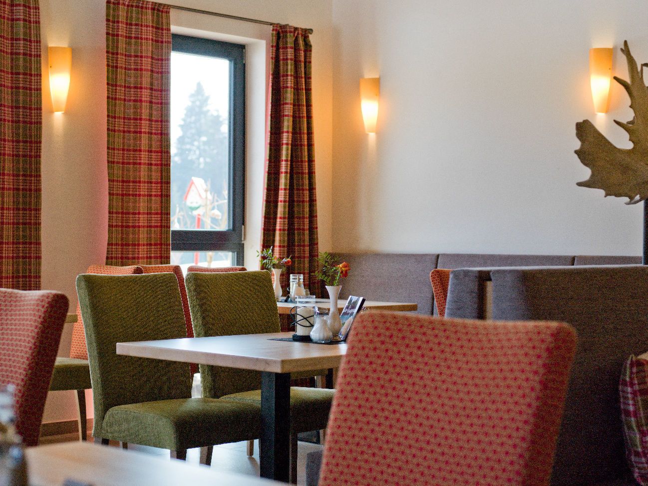 A restaurant interior with plaid curtains, green and red upholstered chairs, and a table with a vase and a menu.