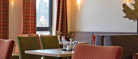 A restaurant interior with plaid curtains, green and red upholstered chairs, and a table with a vase and a menu.