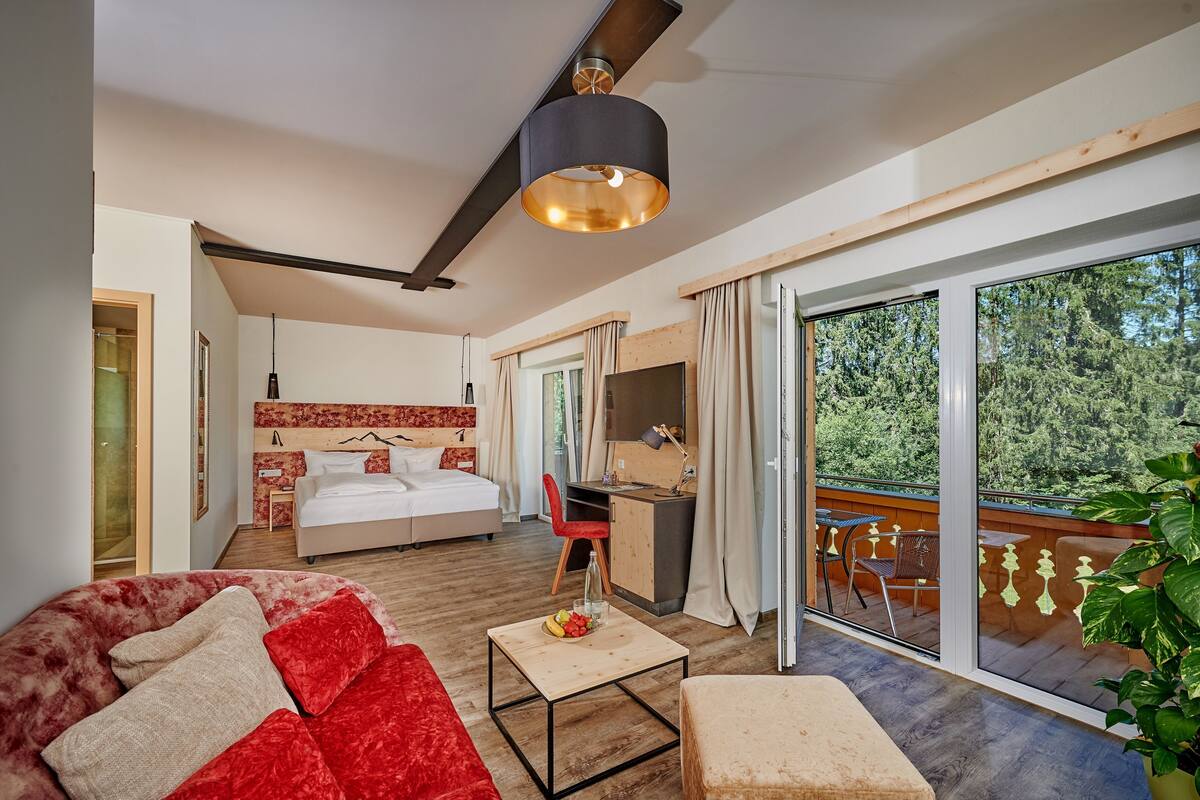 A modern hotel room with a bed, desk, and balcony with a view of trees.
