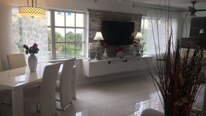 Ein modernes Wohnzimmer mit einem weißen Esstisch, Stühlen, einem Flachbildfernseher und dekorativen Vasen.