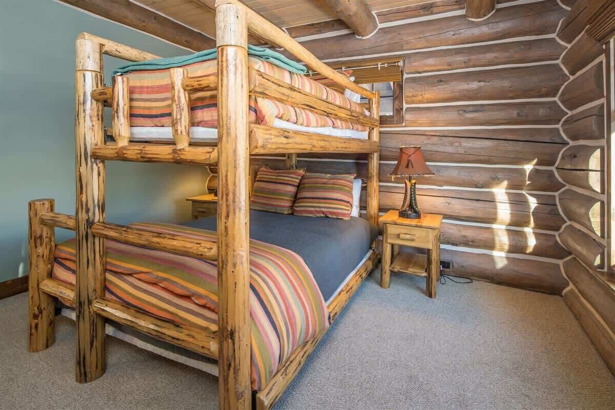 Een stapelbed met strepenbeddengoed, een nachtkastje met een lamp en een wand van houten boomstammen.