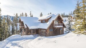 Une cabane en bois, dont le toit est recouvert de neige, entourée d’arbres et située à proximité d’une piste de ski.