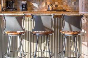 Trois tabourets de bar aux pieds métalliques et aux assises en bois, placés devant un comptoir de cuisine orné d’un dosseret carrelé et surmonté de machines à café.