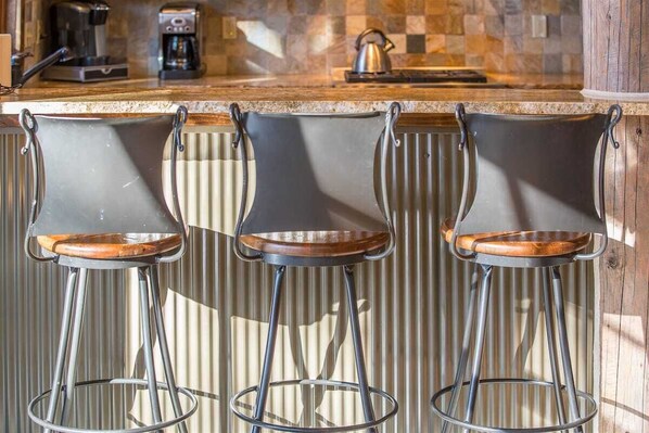Three bar stools with metal legs and wooden seats, positioned in front of a kitchen counter with a checkered tile backsplash and coffee machines.