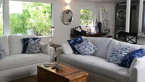 A modern living room with a white sofa, blue patterned pillows, a wooden coffee table, and a large window with a view of greenery. - Eagle Harbor View Cottage (Bainbridge Island)