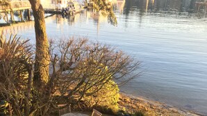 A lakeside patio with a round table and chairs, surrounded by trees and a view of boats on the water. - Eagle Harbor View Cottage (Bainbridge Island)