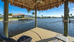 A wooden deck with a thatched roof, overlooking a body of water with a dock and buildings in the background. - Lovely waterfront gulf access, heated pool in SW Cape Coral awaits you. (Cape Coral)