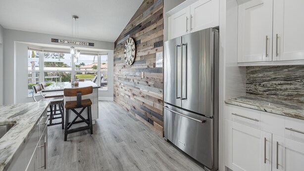 A modern kitchen with a wooden accent wall, a stainless steel refrigerator, and a dining area with a view of the outdoors. - Lovely waterfront gulf access, heated pool in SW Cape Coral awaits you. (Cape Coral)