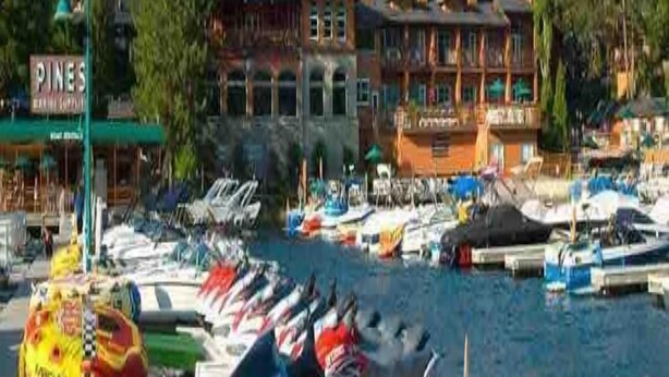 Un port esportiu amb vaixells amarrats, un edifici amb un rètol que diu "Pines" i un vessant forestat al fons.