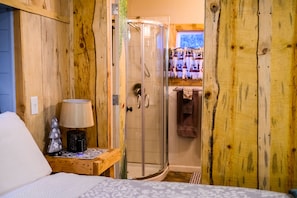 Una habitación de madera con una cama, una mesita de noche con lámpara y un box de ducha con paredes de vidrio.