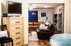 Una habitación con un aparador de madera, un televisor encima, un sofá de cuero marrón, una cama con manta azul y suelo de madera.