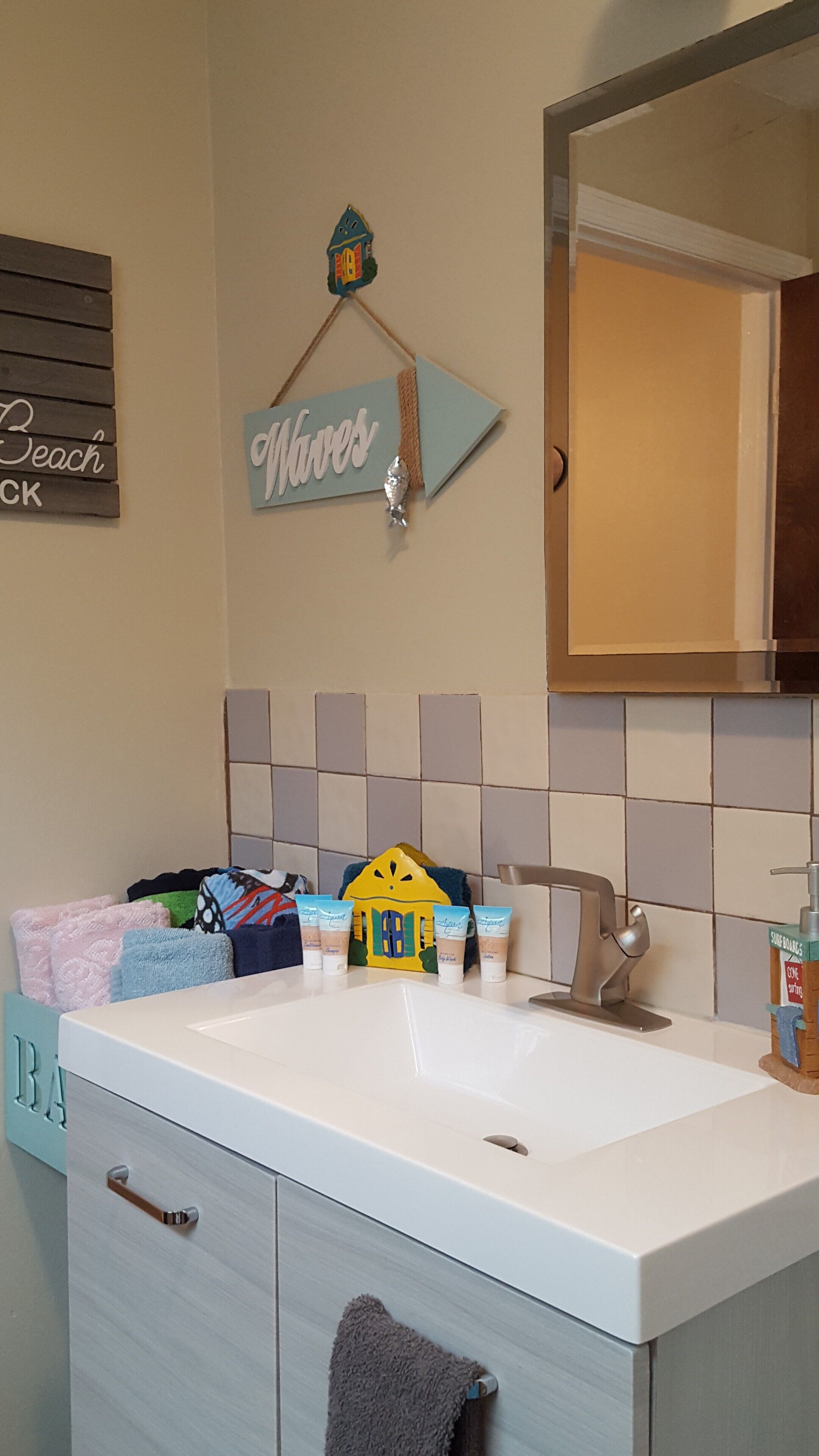 Une salle de bain avec un lavabo blanc, un miroir et un mur recouvert de carreaux. On y trouve un porte-serviettes avec une serviette bleue et un objet jaune qui y est suspendu. Un panneau sur le mur indique : « Plage ».