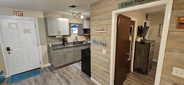 A compact kitchen with a sink, cabinets, and a microwave. There's a sign above the door reading "EXIT" and another sign above the door to the right that says "Beach Place."