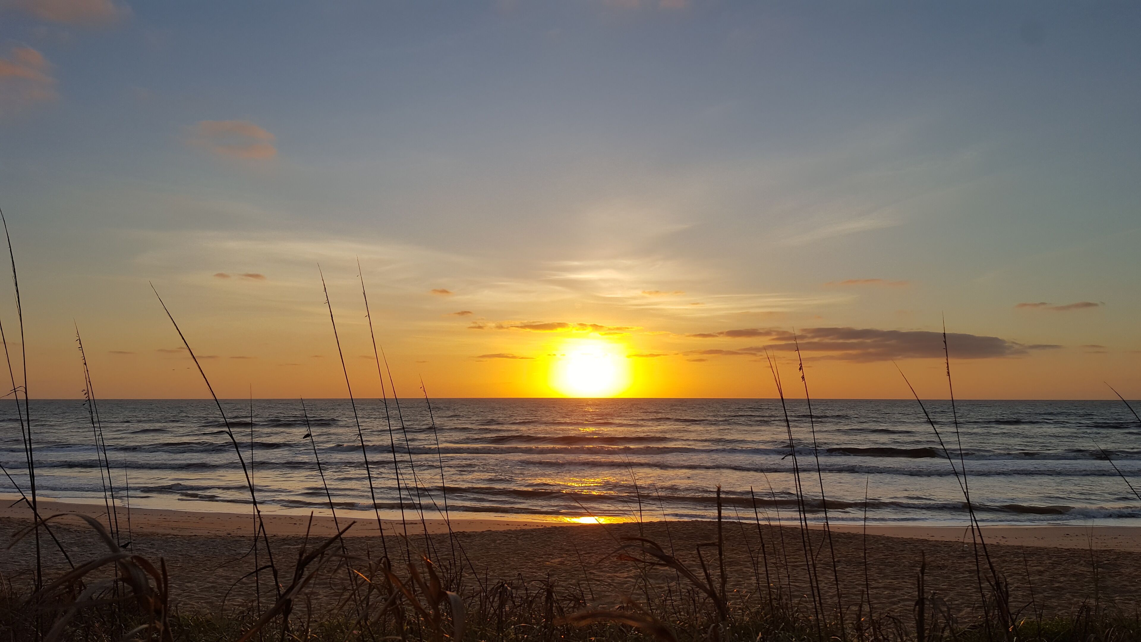 A beach scene with the sun setting over the ocean, waves, and beach grasses.