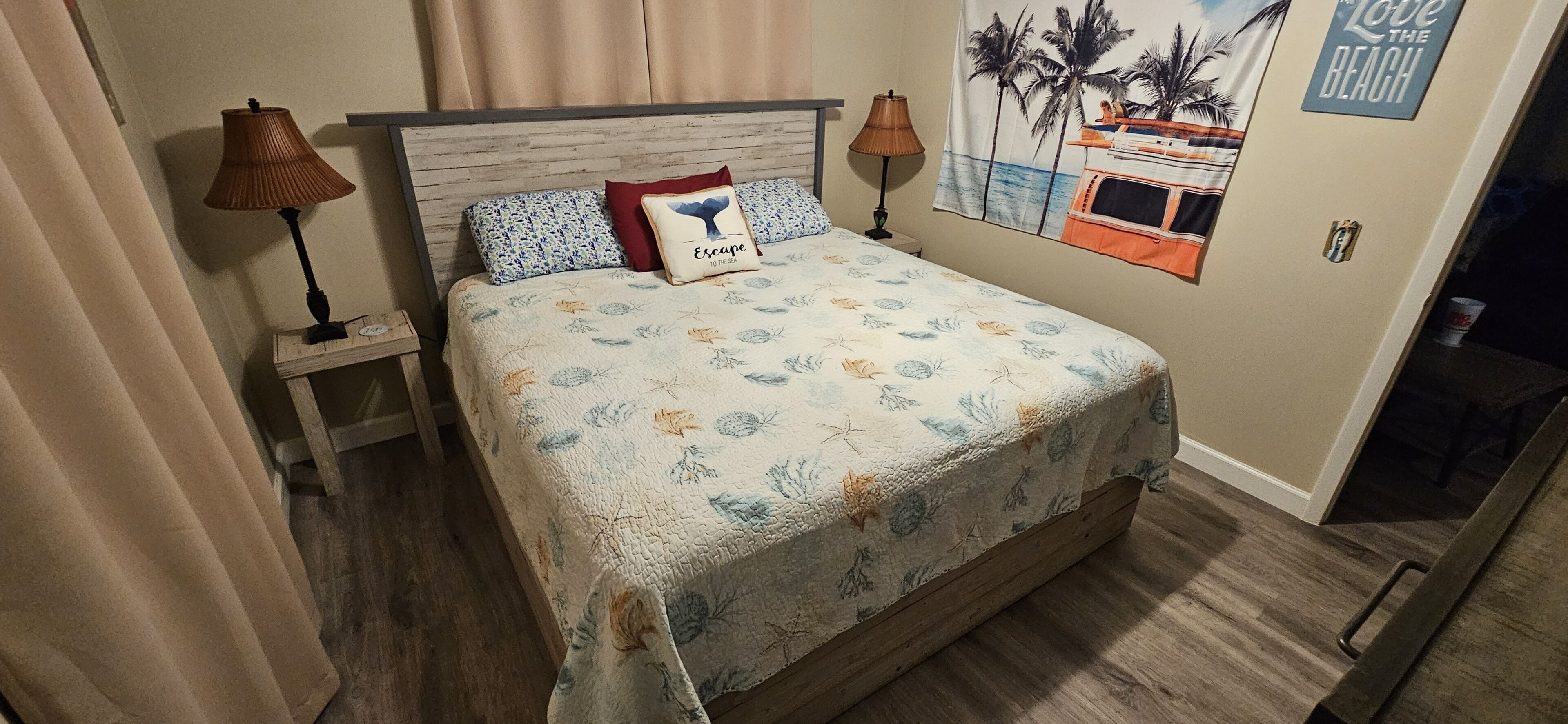Un dormitorio con cabecera de madera, colcha con motivos florales, dos lámparas de noche y un tapiz de pared con temática marina.