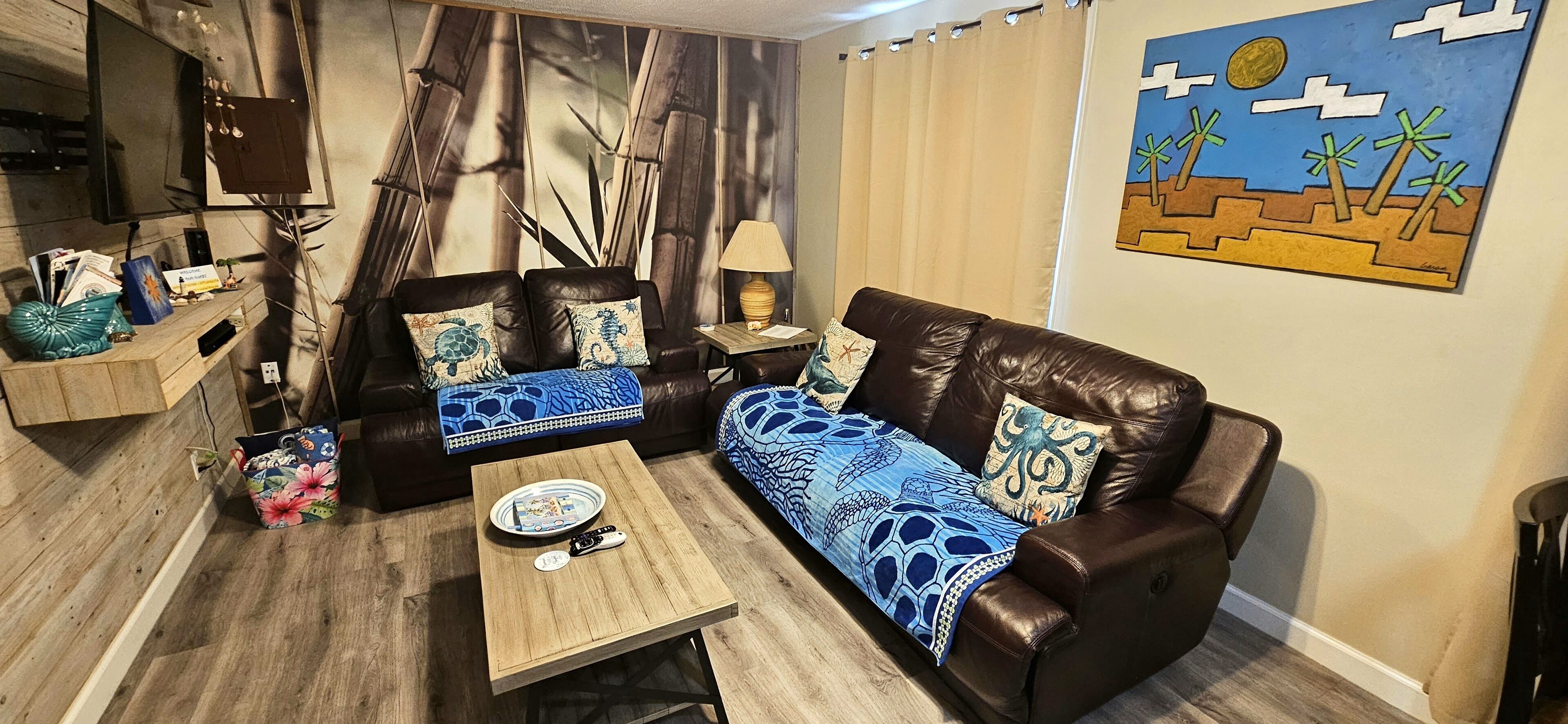 Una sala de estar con un sofá de cuero marrón, una mesa de centro de madera y una pared decorada con un cuadro de temática tropical.