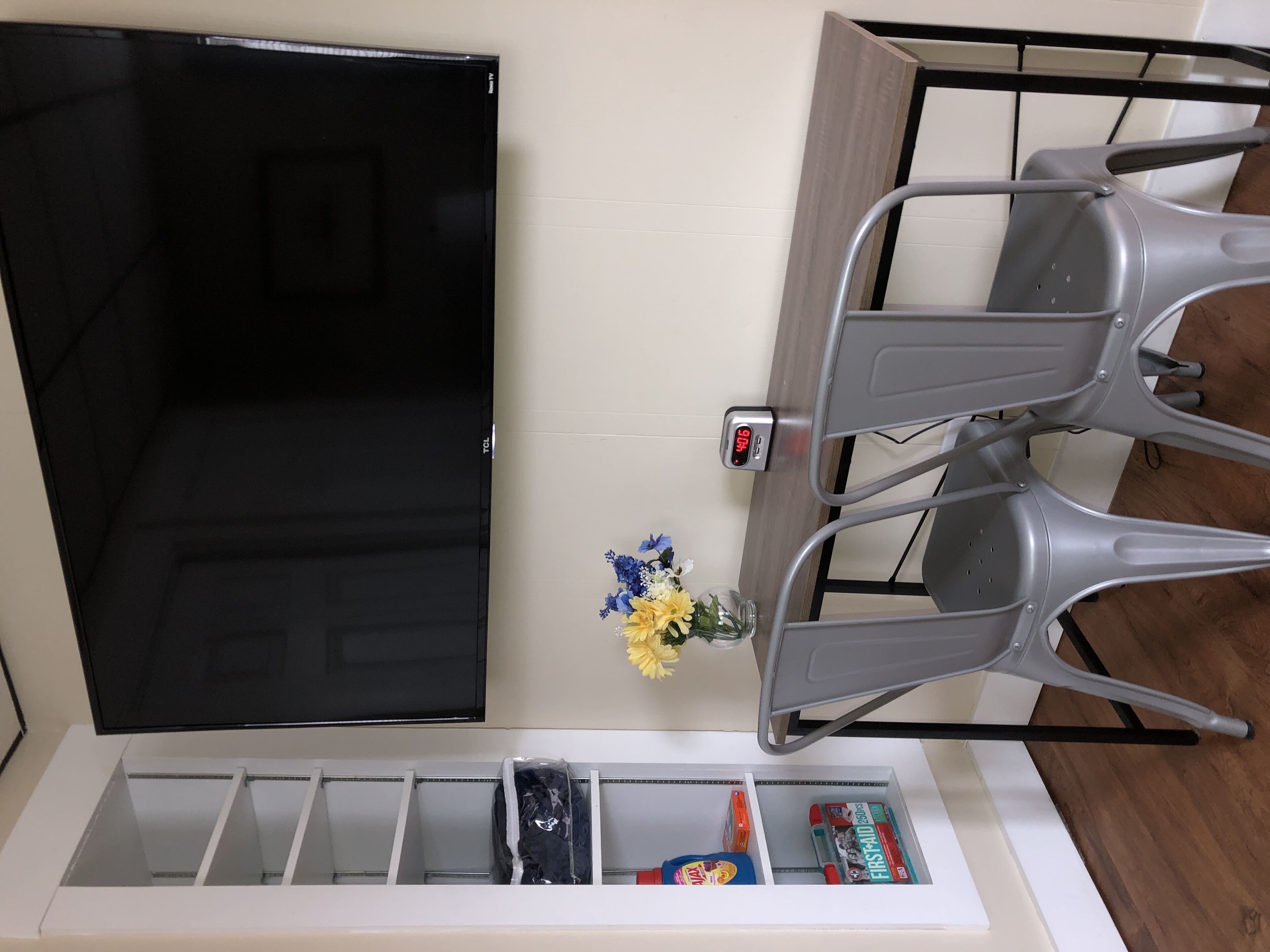 Una habitación con un televisor negro, un estante blanco con diversos objetos y dos sillas grises.