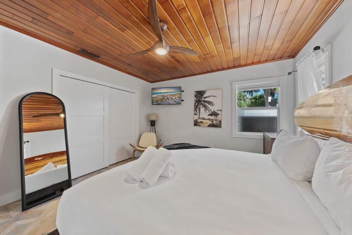 A bedroom with a wooden ceiling, a bed with white linens, a ceiling fan, a window with a view, and a door.