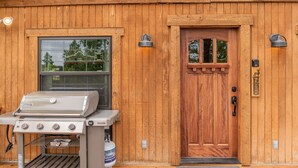 Ein Holzbau mit großem Fenster, einer Tür und einem Willkommensschild, sowie einem Grill vor der Fassade.