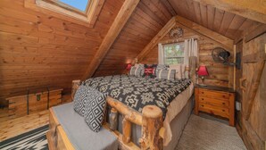 Ein gemütliches Schlafzimmer im Dachgeschoss mit schrägen Wänden, einem Bett mit gemusterten Textilien, einem Nachttisch, einer Lampe und einem Fenster mit Vorhängen.