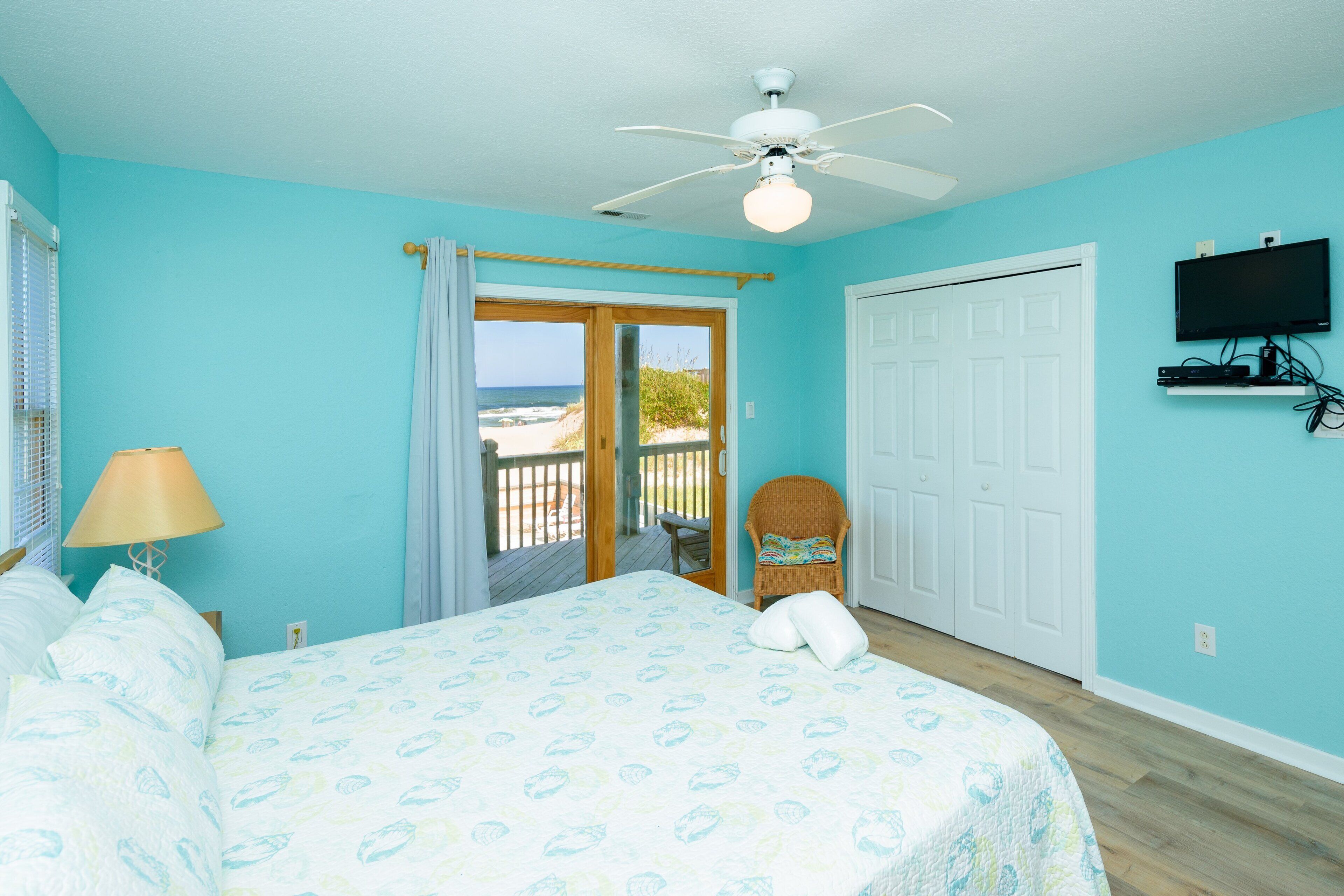 Ein Schlafzimmer mit Bett, Deckenventilator, an der Wand montiertem Fernseher und Blick auf den Strand durch eine Tür.
