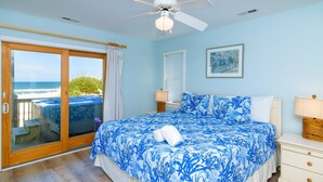 Een slaapkamer met een bed, een plafondventilator, een nachtkastje, een lamp en een raam met uitzicht op het strand.