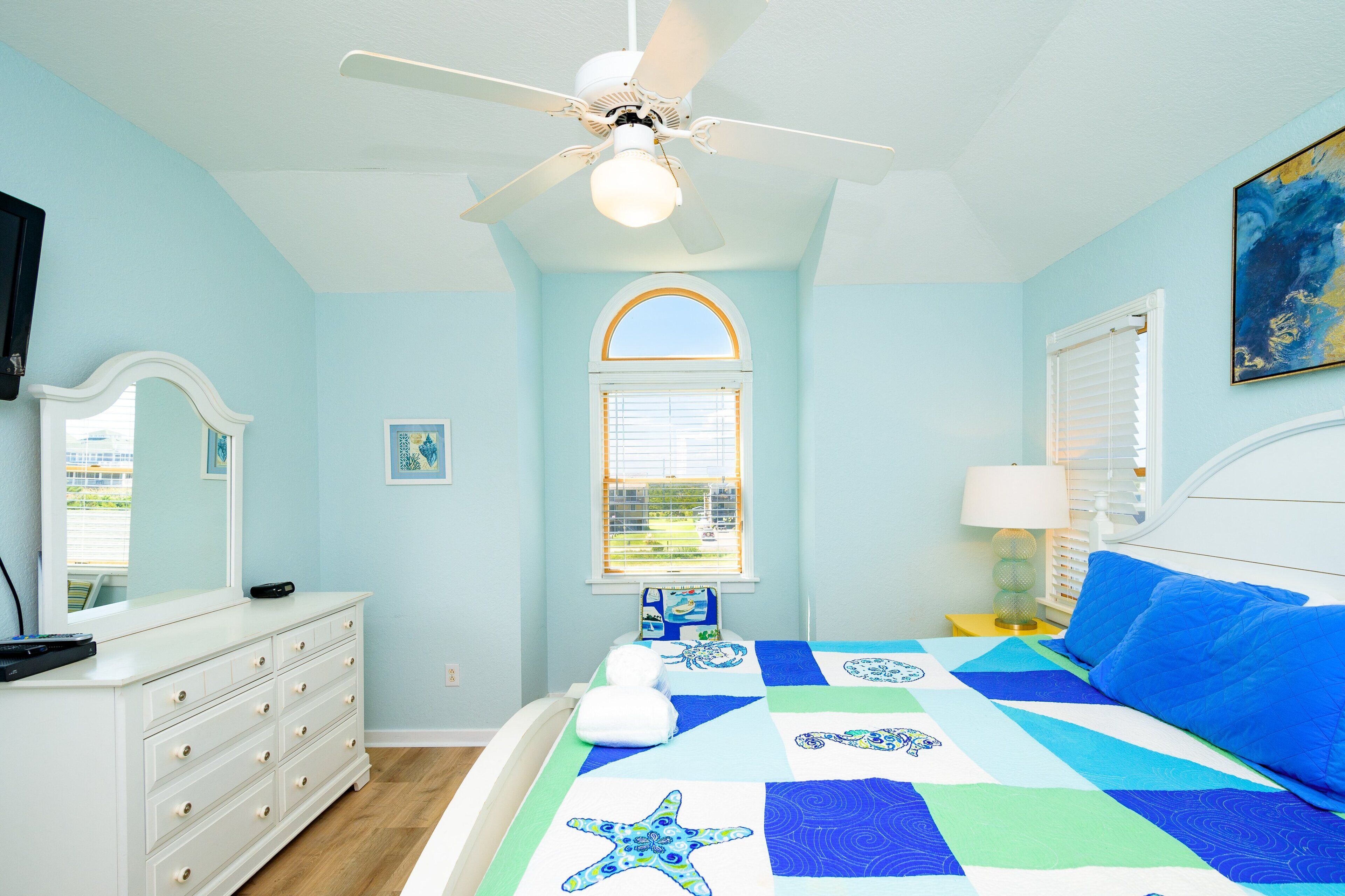 A bedroom with a bed, a dresser, a ceiling fan, a window, and a painting on the wall.