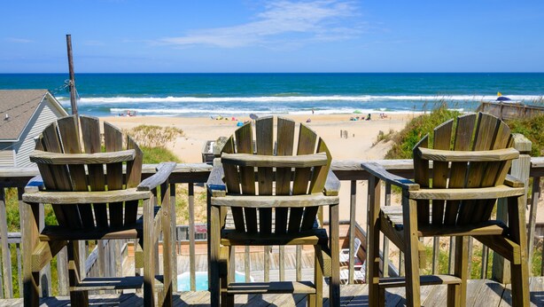 Drie houten Adirondack-stoelen gericht op een strand met een houten terras en uitzicht op de oceaan.