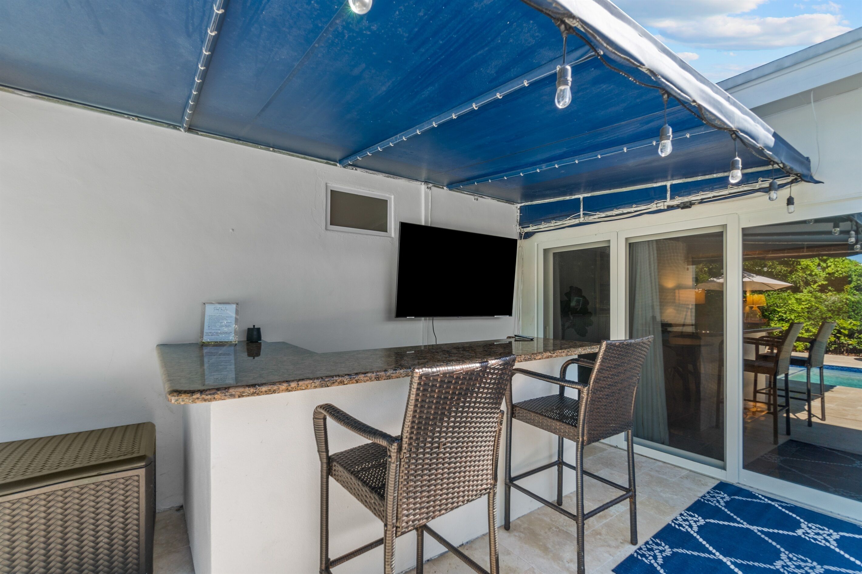 Un área de bar al aire libre con un televisor de pantalla plana, dos sillas de mimbre, una barra y un toldo azul.